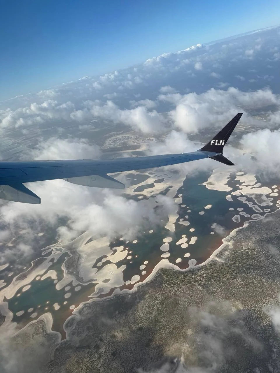 Крыло самолёта с надписью FIJI над облаками, водой и светлыми отмелями